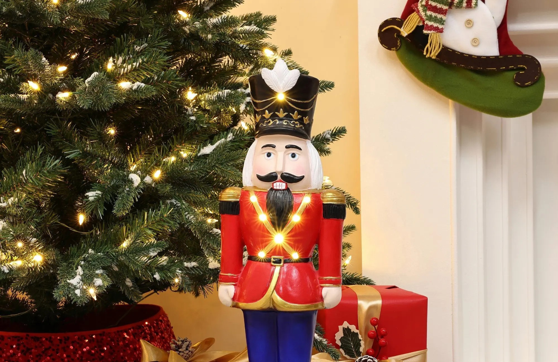 A nutcracker figurine in a red uniform stands in front of a decorated Christmas tree and a wrapped gift.