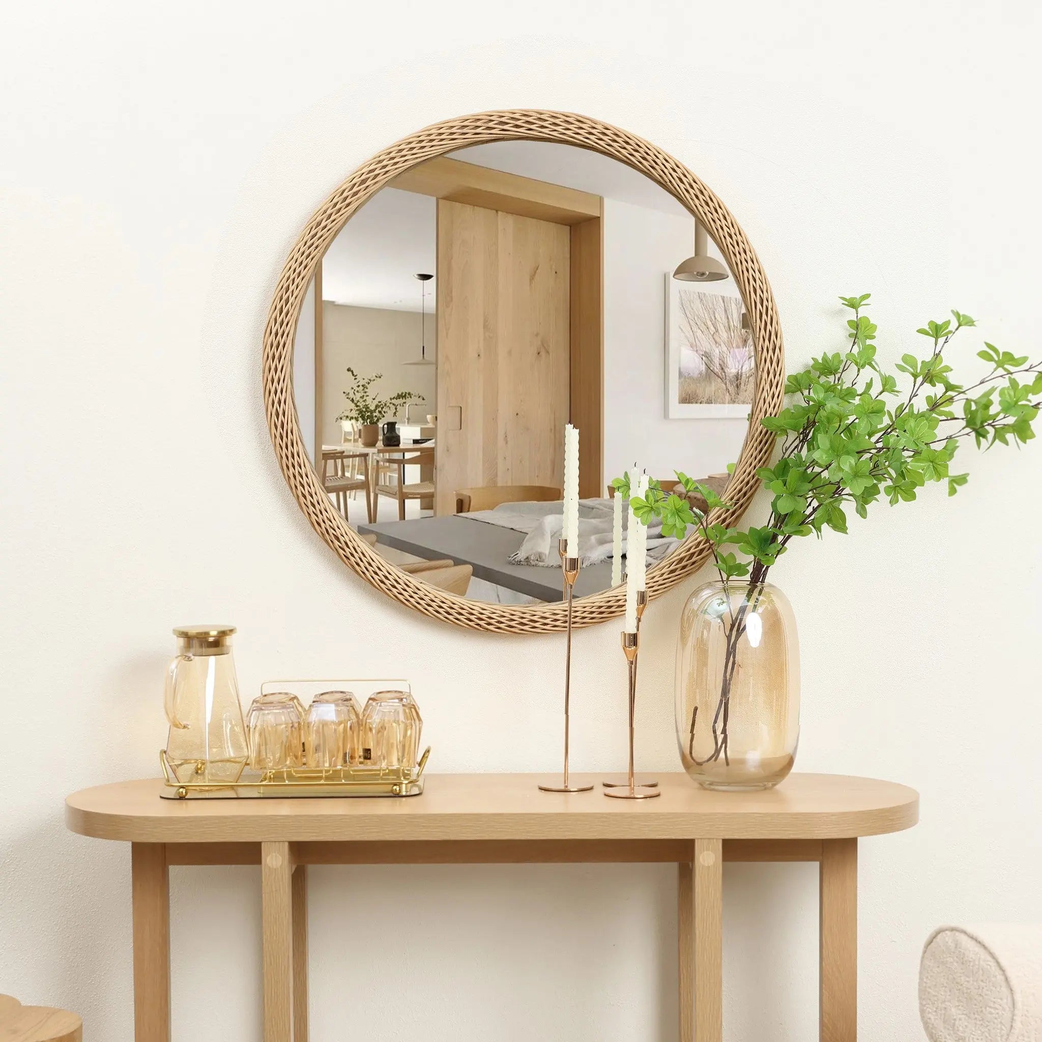 A decorative mirror with a circular design hangs above a wooden console table. On the table sit a vase with leafy branches, two tall candlesticks, and a tray with a pitcher and glasses, creating an inviting tableau.