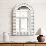 The stylish home decor features an arched rectangular design mirror with a ribbed frame on a white brick wall, reflecting a window; a vase and lamp sit elegantly on the wooden dresser below.