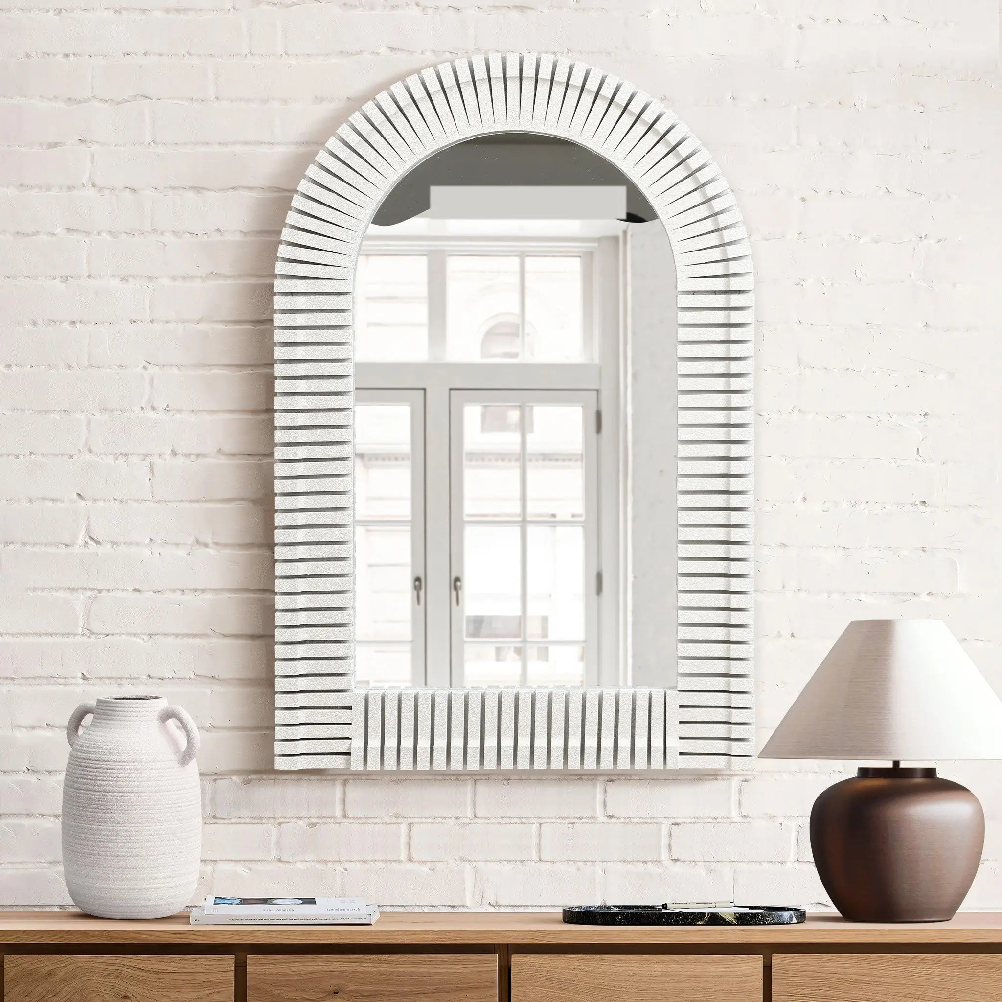 The stylish home decor features an arched rectangular design mirror with a ribbed frame on a white brick wall, reflecting a window; a vase and lamp sit elegantly on the wooden dresser below.