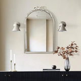 A modern romantic style is evident in the decorative Ribbon Wall Mirror, showcasing an arched silhouette and elegant bow detail above a dark console table. Two candles and a vase with branches accompany the mirror, while cone-shaped wall sconces gracefully flank it.
