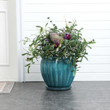 A green ceramic decorative planter, adorned with lush foliage and purple flowers, sits gracefully on a gray floor in front of a white paneled wall.
