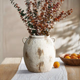 A terracotta jug vase with dried eucalyptus branches sits on a wooden table, exuding vintage appeal, next to a handcrafted wooden bowl filled with apricots.