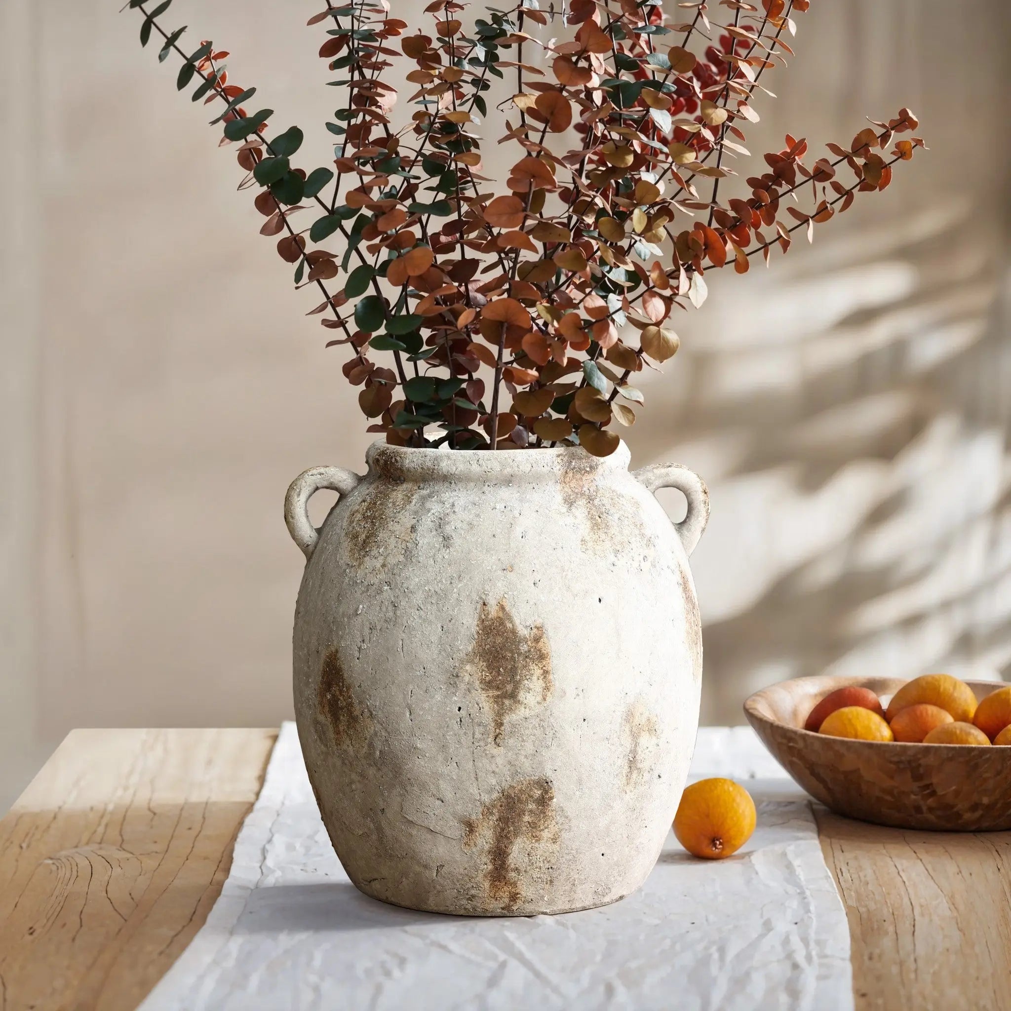 A terracotta jug vase with dried eucalyptus branches sits on a wooden table, exuding vintage appeal, next to a handcrafted wooden bowl filled with apricots.