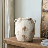A handcrafted, rustic terracotta jug vase with handles sits on a wooden table next to a framed artwork, exuding vintage appeal.