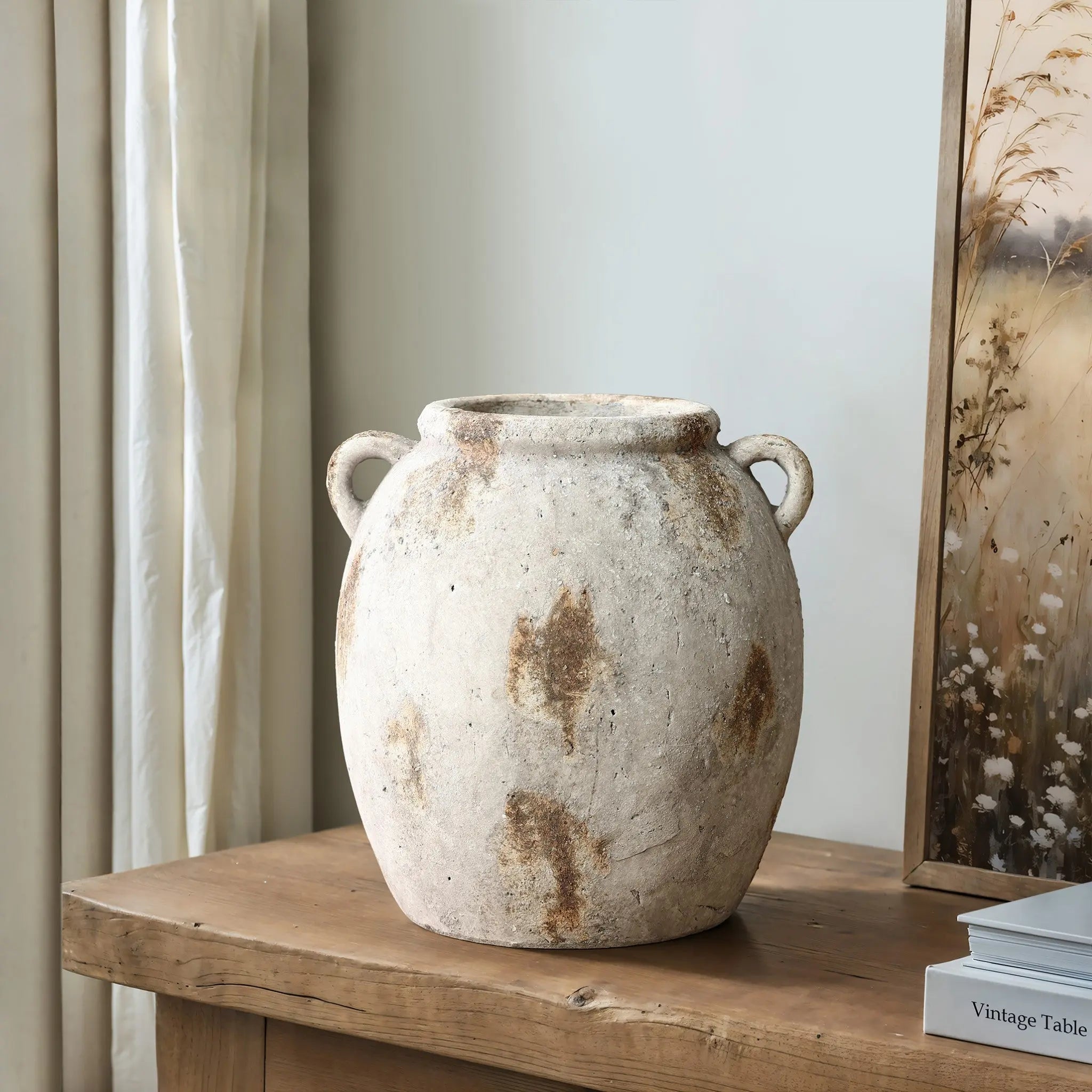 A handcrafted, rustic terracotta jug vase with handles sits on a wooden table next to a framed artwork, exuding vintage appeal.