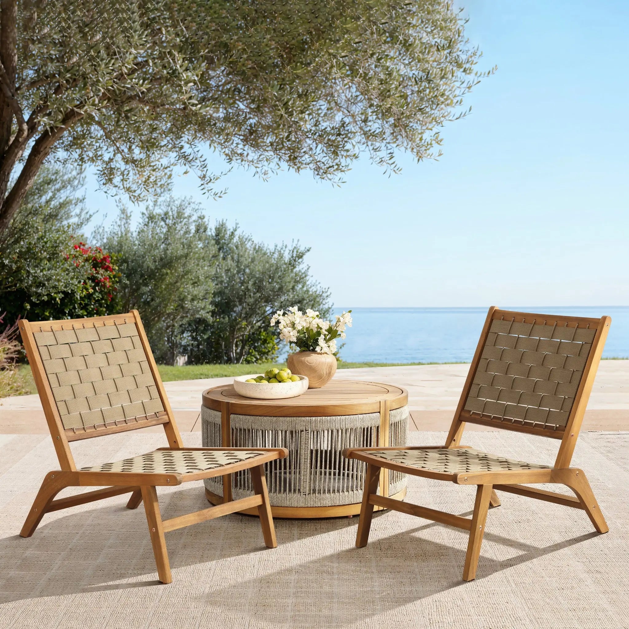 Two woven chairs crafted from acacia wood face each other across a round table with a fringe. A vase with flowers and a bowl sit atop the outdoor furniture. Olive trees and a sea view paint the serene background.