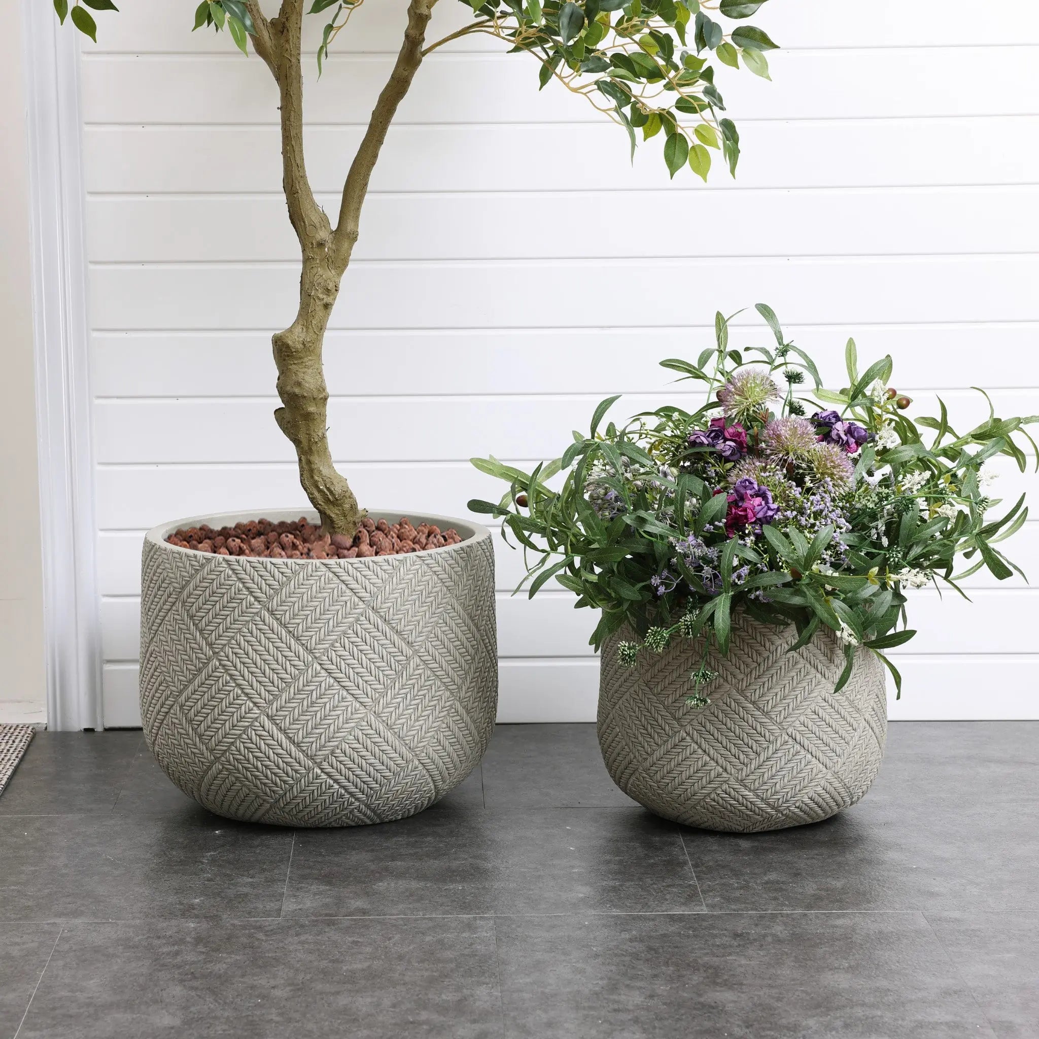 Two textured planters on a tiled floor, crafted from durable MgO material—one cradling a leafy tree and the other brimming with wildflowers and greenery. A white-paneled wall stands in the background, enhancing their charm.