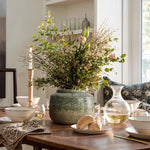 A dining table features a Rustic brown stoneware bowl vase filled with leafy branches and small flowers. It is surrounded by white plates, bowls, a tall candle holder, and a clear glass pitcher. A patterned cushion adorns a chair in the background, adding elegance to the home decor ambiance by LuxenHome.
