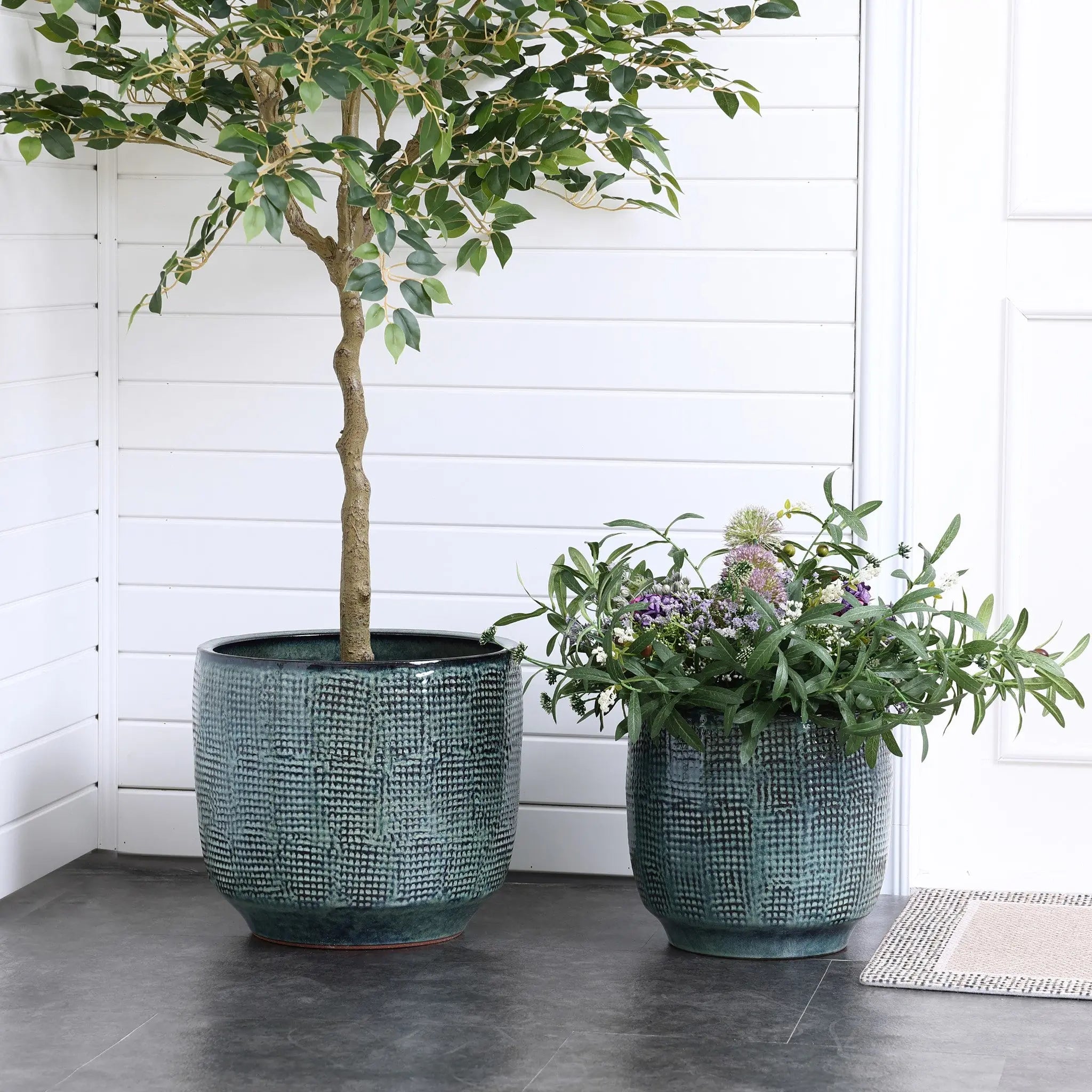Two Marina gray glazed round ceramic planters by LuxenHome sit on a tiled floor. One holds a lush, leafy tree while the other features flowering plants. The setting includes white wooden paneled walls and a doormat near a door.