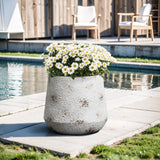 A large gray MgO material planter with white daisies sits gracefully on a tiled patio near the swimming pool, exuding a vintage minimalist style. In the background, two wooden chairs and a table complete the serene setting.