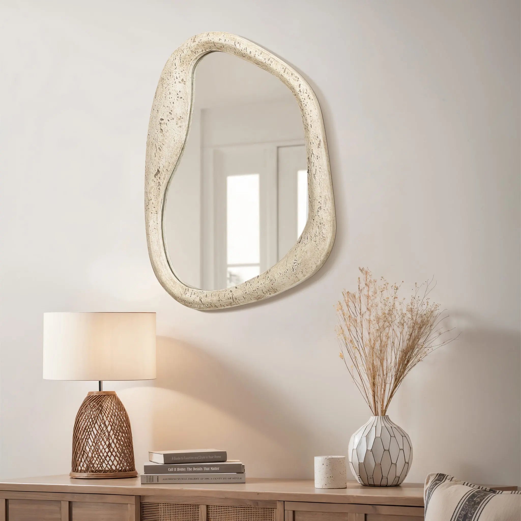 As an eye-catching piece of home decor, this asymmetrical mirror with a free-form frame hangs above a wooden console. Positioned elegantly beside a lamp and a vase with dried flowers, its unique design adds charm and sophistication to any space.