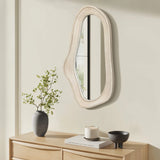 An oval irregularly shaped wall mirror with a natural finish wood frame hangs above a wooden dresser. Its adorned with a small plant, lit candle, and black bowl, embodying the essence of modern home decor.