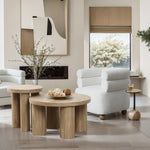 A modern living room features an oak-finish coffee table, a white cushioned chair, abstract wall art, and a view of trees through the window. The decor includes vases with branches and decorative spheres for a serene vibe.
