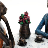 Clay figures, reminiscent of romantic moments, are seated around a table with coffee cups and a vase of red roses in the center. This charming scene makes for a delightful wedding gift or modern brown resin couple decoration.