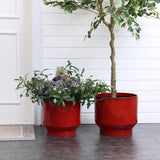 Two burgundy red glazed round ceramic planters from LuxenHome rest on a tiled floor by a white paneled wall. One, with lush greenery and small purple flowers, features vertical stripes; the other contains a small tree with light green leaves. A doormat appears partially on the left.