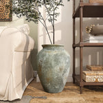 A Rustic gold and brown terracotta urn vase by LuxenHome, 15.2 h, stands on a wooden floor beside a beige sofa, holding tall branches with delicate leaves. Nearby, a brown shelf filled with books and decor items enhances the cozy, natural ambiance using an antique distressing technique.