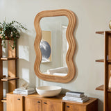 A wavy-edged wooden mirror adds a modern vibe as it hangs above a wooden cabinet with a white bowl and stacked books. A plant sits on the cabinet, while cork wood details enhance the aesthetic. A framed artwork is partially visible in the mirrors reflection.