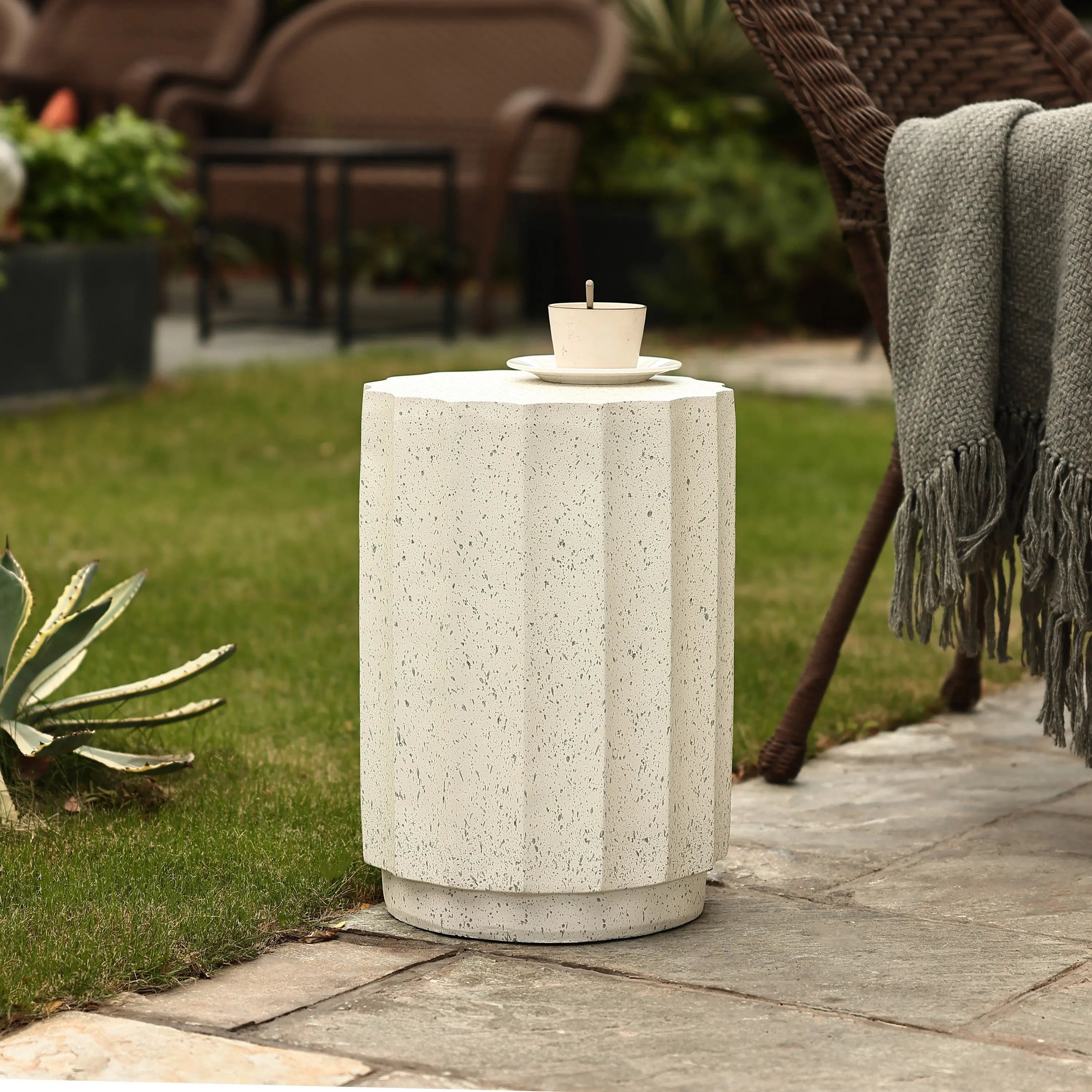 A white, textured cement modern side table with a cup and saucer sits on a patio next to a wicker chair and green lawn, adding style to your outdoor decor.