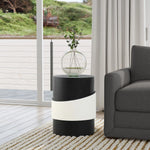 A contemporary living room showcases a black side table with a white band and robust metal construction. A round glass vase with green branches graces the table. In the background, a gray sofa and large window with crisp white curtains complete the modern minimalist design.
