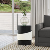 A contemporary living room showcases a black side table with a white band and robust metal construction. A round glass vase with green branches graces the table. In the background, a gray sofa and large window with crisp white curtains complete the modern minimalist design.