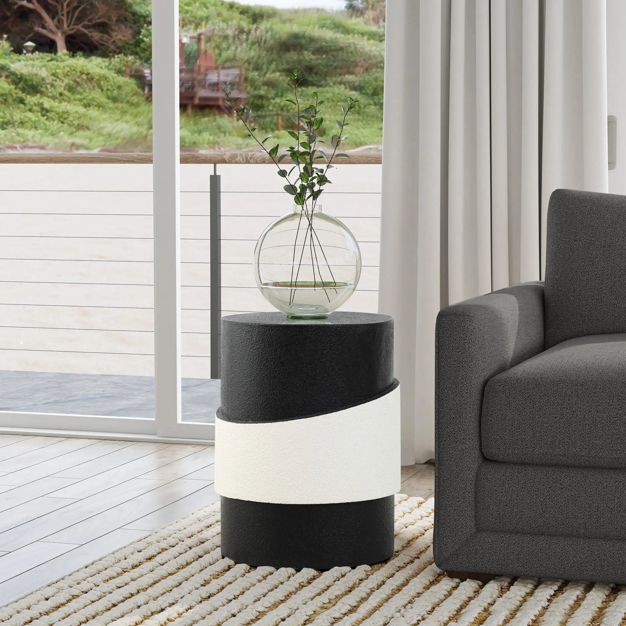 A contemporary living room showcases a black side table with a white band and robust metal construction. A round glass vase with green branches graces the table. In the background, a gray sofa and large window with crisp white curtains complete the modern minimalist design.
