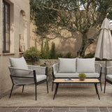 The outdoor patio features a cozy seating arrangement with an outdoor aluminum rattan sofa and chairs, all adorned with Olefin fabric cushions. A small table, topped with a potted plant, sits shaded under an umbrella amid lush plants in the background.