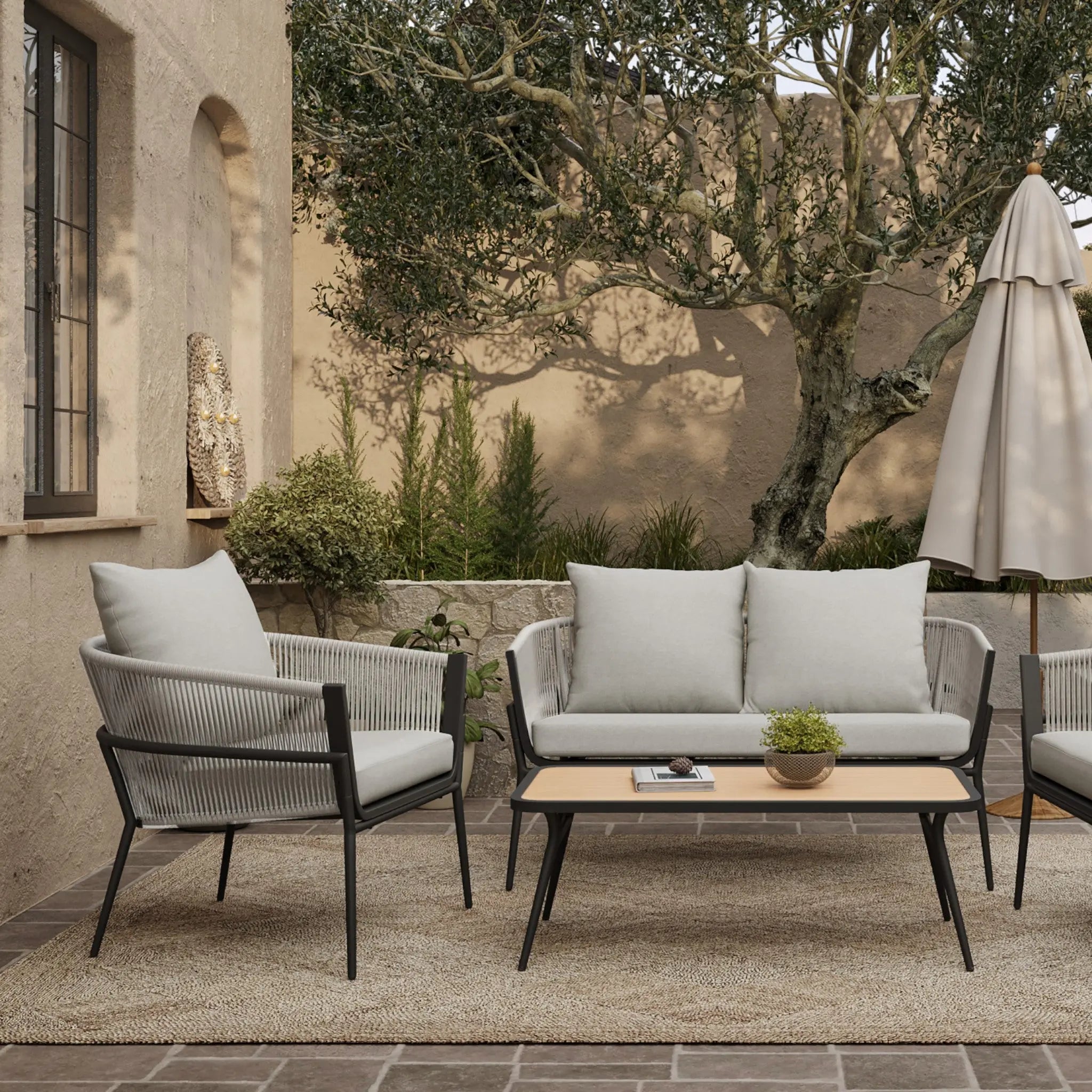 The outdoor patio features a cozy seating arrangement with an outdoor aluminum rattan sofa and chairs, all adorned with Olefin fabric cushions. A small table, topped with a potted plant, sits shaded under an umbrella amid lush plants in the background.