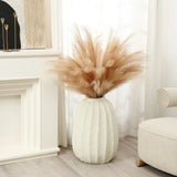 A modern white vase filled with tan pampas grass is placed on a wooden floor next to a white fireplace and a cream chair, serving as an artistic centerpiece in this minimalist home decor setting, with curtains gracefully framing the background.