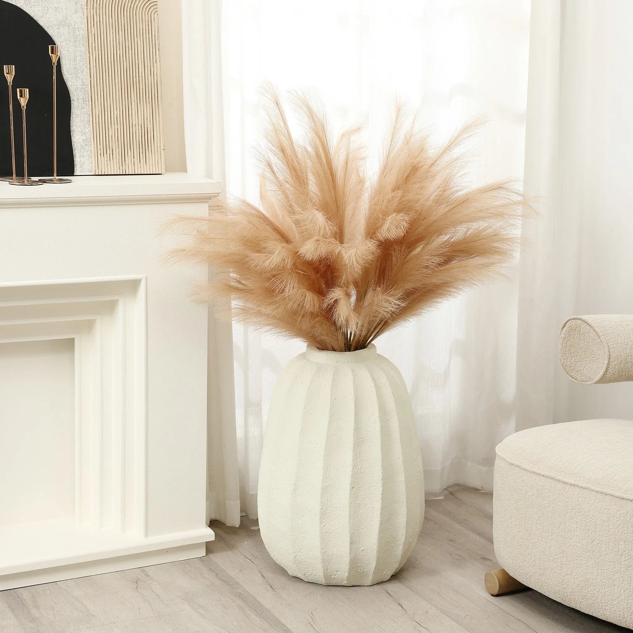 A modern white vase filled with tan pampas grass is placed on a wooden floor next to a white fireplace and a cream chair, serving as an artistic centerpiece in this minimalist home decor setting, with curtains gracefully framing the background.