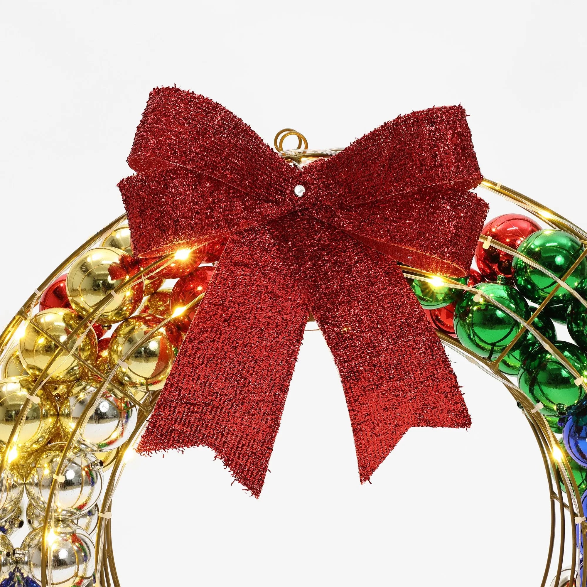 A festive holiday wreath with a large red glittery bow, decorated with gold, red, green, and blue ornaments. Perfect as an outdoor holiday decoration, its enhanced with twinkling LED lights for an extra touch of seasonal magic.