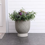 A large MgO material planter with an intricate pattern holds a variety of purple and white flowers with lush green foliage, elegantly placed on a dark tiled floor against a white paneled wall.