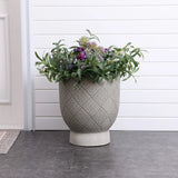 A large MgO material planter with an intricate pattern holds a variety of purple and white flowers with lush green foliage, elegantly placed on a dark tiled floor against a white paneled wall.