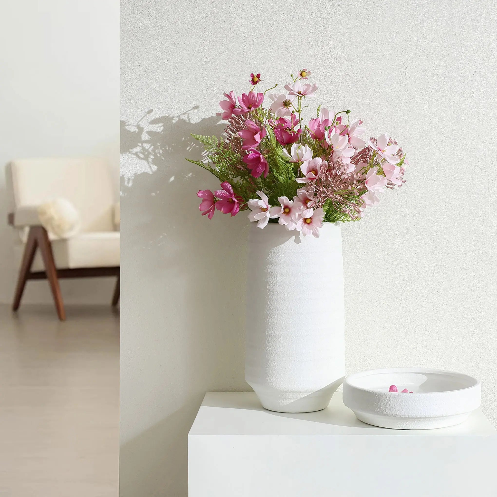 A white terracotta vase adorned with pink and white flowers graces a white table. Beside it rests a shallow dish, capturing the simplicity of the scene. A beige armchair completes this tranquil indoor setting, offering a cozy retreat.