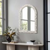A vintage wall mirror with an ornate white frame hangs on a gray wall above a light wood console table. A black vase with yellow flowers and a stack of books sit elegantly on the table, while the windows light reflects beautifully in the arched mirror.