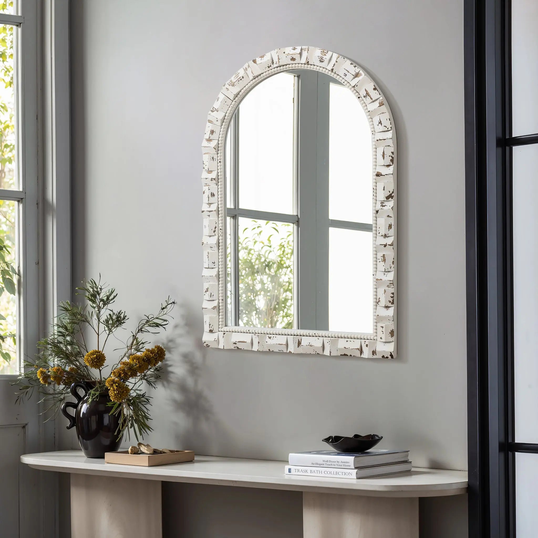 A vintage wall mirror with an ornate white frame hangs on a gray wall above a light wood console table. A black vase with yellow flowers and a stack of books sit elegantly on the table, while the windows light reflects beautifully in the arched mirror.