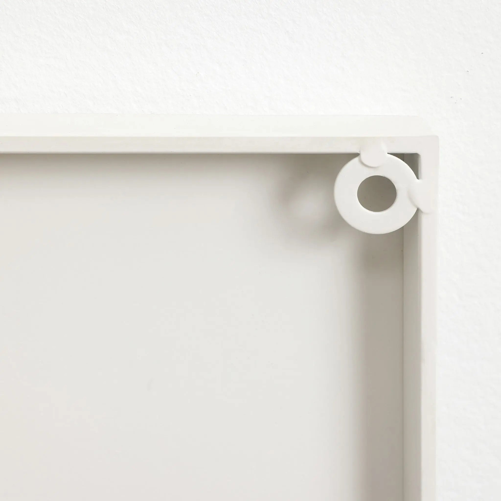 Close-up of a light gray corner of a rectangular frame with a round, white hook attached, reminiscent of abstract metal wall decor on a pristine white wall.