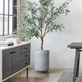 Tall potted plant with slender green leaves in a Gray Round Planter, beside a modern black cabinet and near a dark wooden table with metal legs, all set against the backdrop of large windows for that perfect touch of modern minimalist decor.