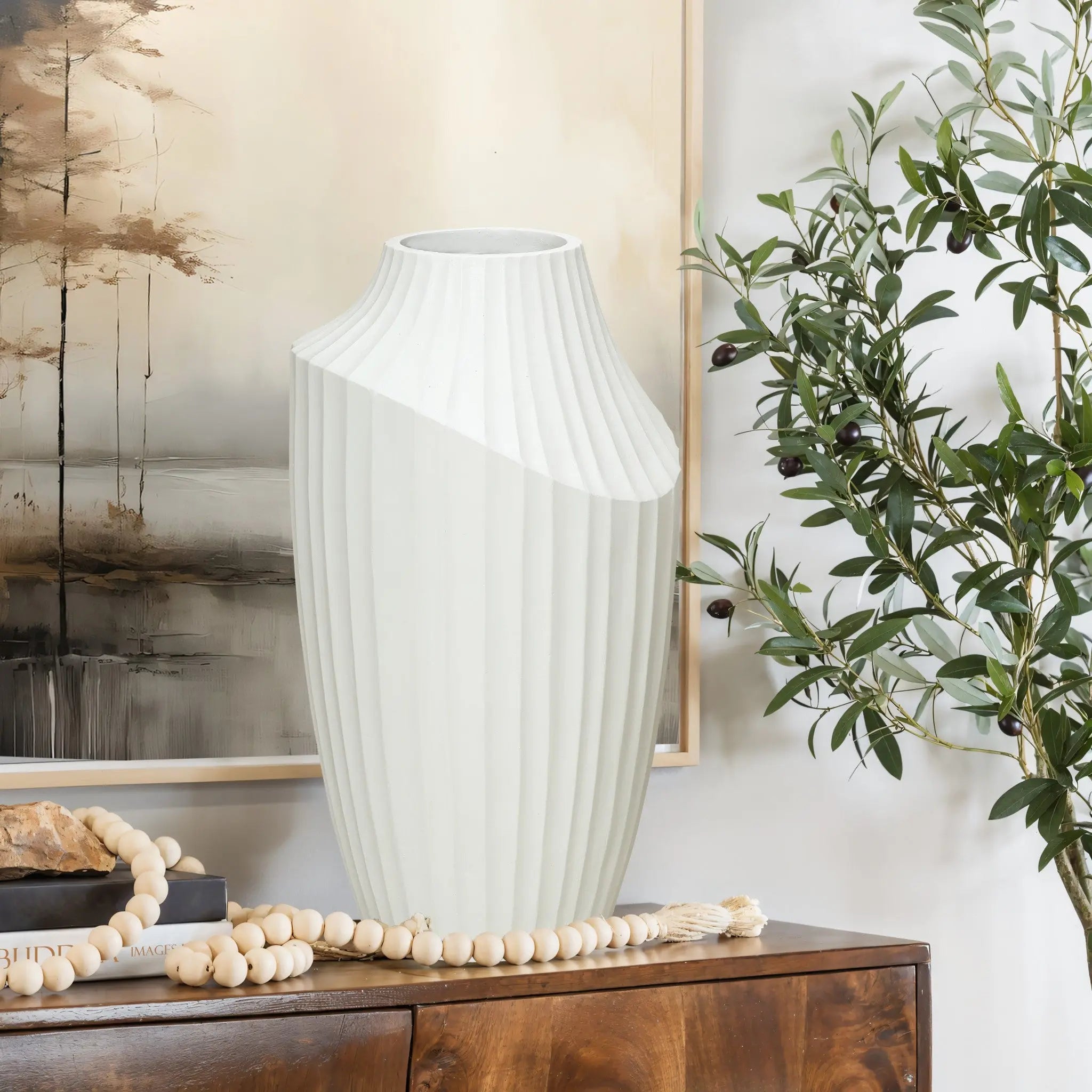 A large, white, vertically ribbed vase serves as a modern abstract sculpture on a wooden table adorned with decorative beads. An abstract painting and an olive tree branch in the background complete the look of contemporary home decor.
