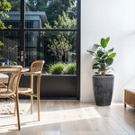 A bright, modern dining area features wooden chairs around a table and a LuxenHome Sea Gray Ripples Glazed Tall Ceramic Planter with a large potted plant on the right. Glass doors reveal the garden, flooding sunlight onto the light wooden floor and minimalist decor with black-and-white striped accents.