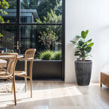 A bright, modern dining area features wooden chairs around a table and a LuxenHome Sea Gray Ripples Glazed Tall Ceramic Planter with a large potted plant on the right. Glass doors reveal the garden, flooding sunlight onto the light wooden floor and minimalist decor with black-and-white striped accents.