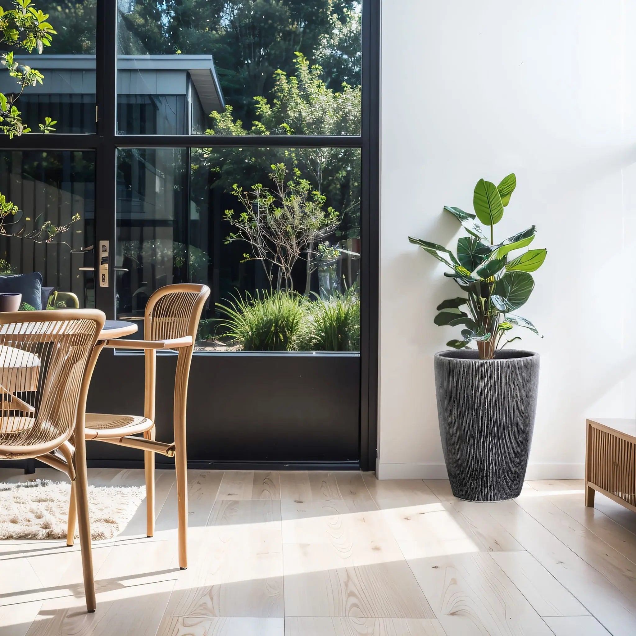 A bright, modern dining area features wooden chairs around a table and a LuxenHome Sea Gray Ripples Glazed Tall Ceramic Planter with a large potted plant on the right. Glass doors reveal the garden, flooding sunlight onto the light wooden floor and minimalist decor with black-and-white striped accents.
