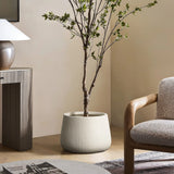 A minimalist living room features a LuxenHome Ribbed Round Planter (10.63 h) on the wooden floor, a textured armchair, and a round table with a white lamp and magazines, creating modern minimalist decor.