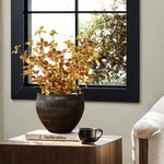 A cozy room showcases a wooden table adorned with a LuxenHome dark brown textured resin round planter filled with yellow and orange foliage. Nearby, the book Authentics and a black mug complement the scene, while a large window offers views of green foliage outside.