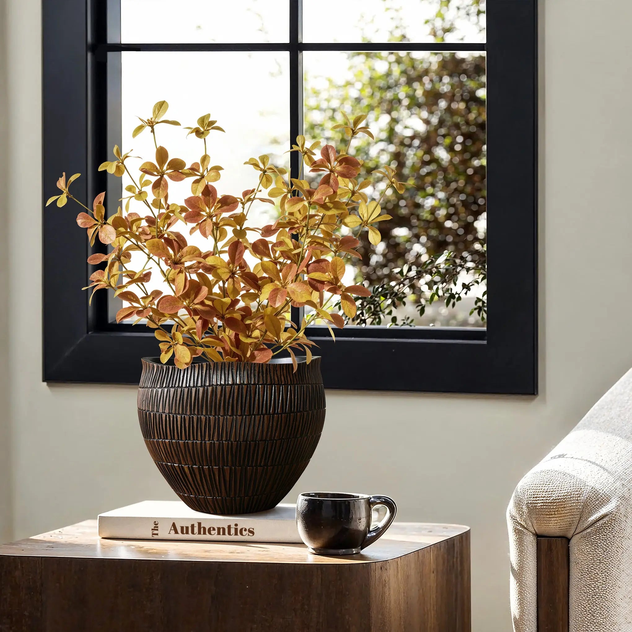A cozy room showcases a wooden table adorned with a LuxenHome dark brown textured resin round planter filled with yellow and orange foliage. Nearby, the book Authentics and a black mug complement the scene, while a large window offers views of green foliage outside.