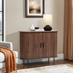 A modern living room showcases the LuxenHome Aria 2-door tambour accent cabinet in a dark oak finish, topped with a lamp, white vase, and black bowl. A framed abstract painting hangs above, an orange throw blanket drapes over the sofa, while beige curtains and a light rug complete the scene.