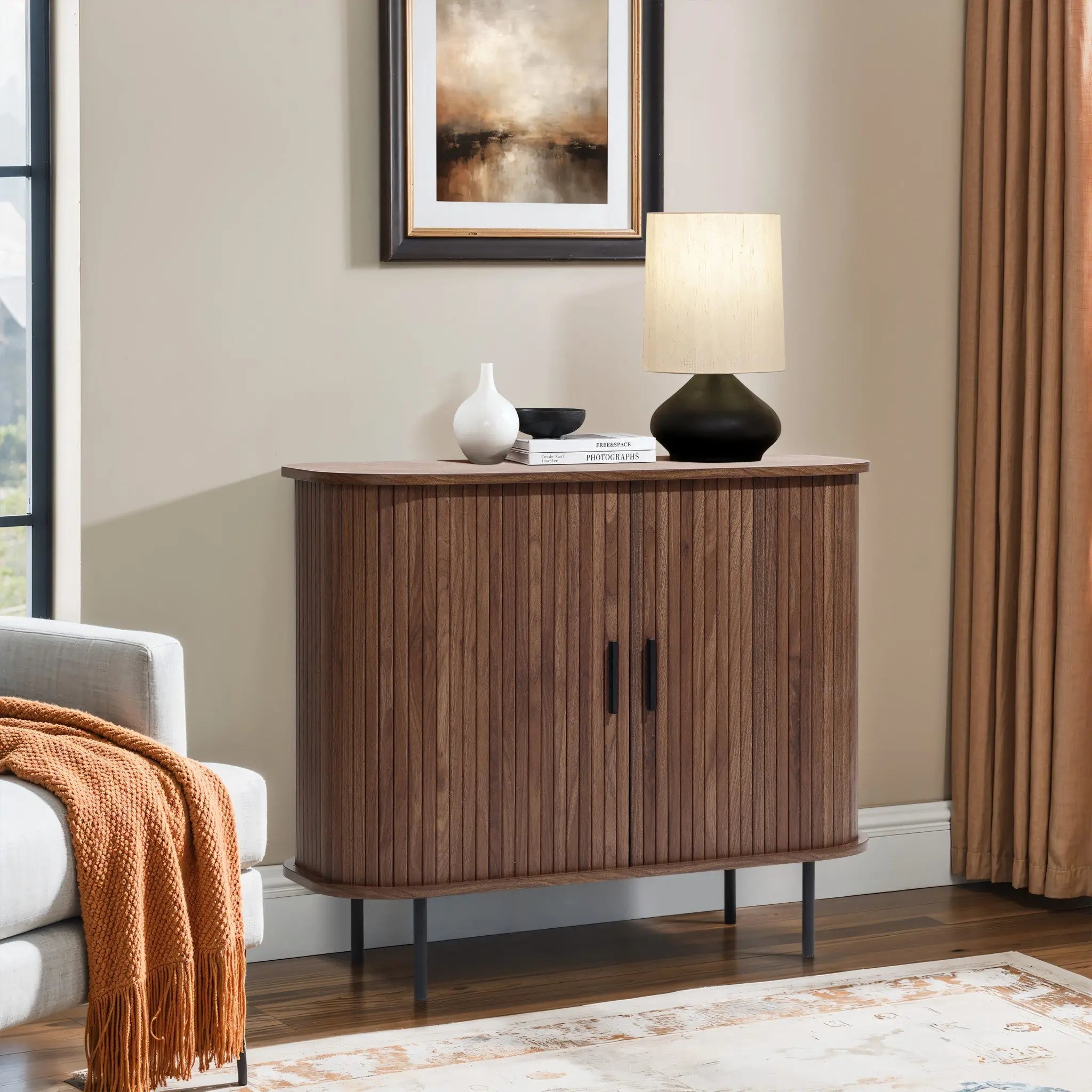 A modern living room showcases the LuxenHome Aria 2-door tambour accent cabinet in a dark oak finish, topped with a lamp, white vase, and black bowl. A framed abstract painting hangs above, an orange throw blanket drapes over the sofa, while beige curtains and a light rug complete the scene.