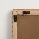 The back of a decorative wall mirror showcases a natural beech wood frame with scalloped edges against a white wall, featuring metal hooks and brackets for hanging.