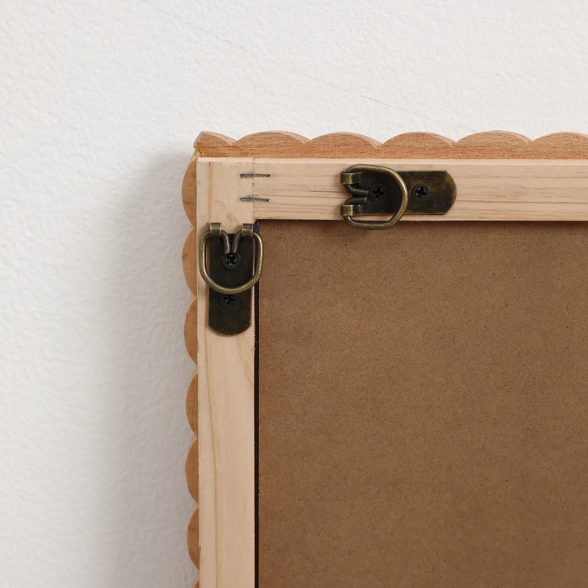 The back of a decorative wall mirror showcases a natural beech wood frame with scalloped edges against a white wall, featuring metal hooks and brackets for hanging.