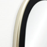 Close-up of an oblong-shaped, contemporary mirrors edge with a striking gold and black frame against a pristine white wall.